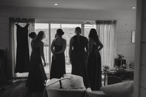 Black and white photo of bridesmaids silhouetted against a bright window, wearing elegant gowns during wedding preparations.