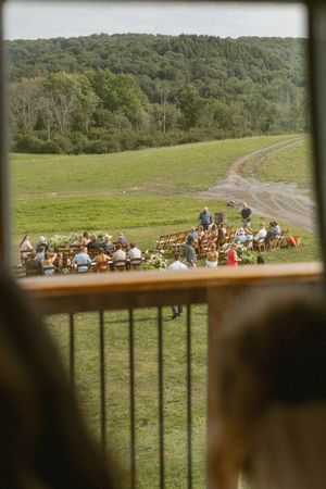 Outdoor wedding ceremony viewed through barn window, guests seated in chairs on green hillside with wooded backdrop.