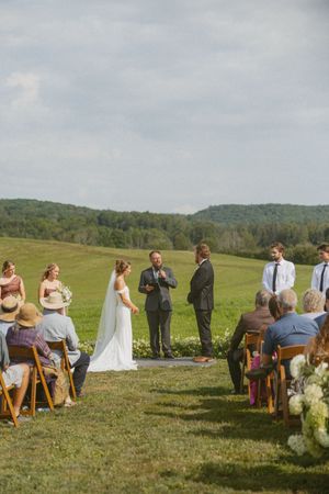 Outdoor wedding ceremony in a green field with bride, groom, and officiant standing at the altar as guests watch.