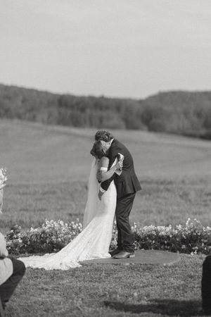 Black and white photo of a bride and groom sharing their first kiss at an outdoor wedding ceremony in a scenic field.