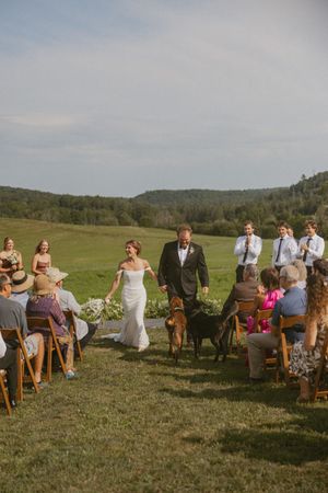 Bride and groom walk down outdoor aisle with dogs at rustic countryside wedding ceremony.