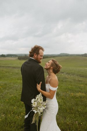 Bride and groom share romantic moment in green meadow, bride holding white floral bouquet.