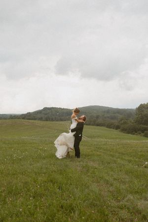 Groom lifting bride in white dress in lush green meadow with rolling hills and overcast sky on wedding day.