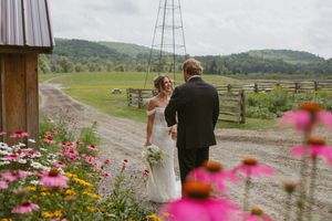 Bride and groom hold hands at a rustic farm wedding with colorful wildflowers and scenic green hills in the background.