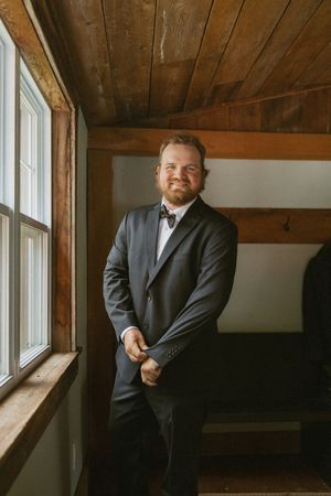 Groom in dark suit and bow tie smiling by rustic wooden window before wedding ceremony.