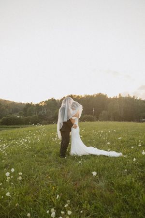 Bride and groom share a romantic kiss under a flowing veil in a sunlit wildflower meadow at sunset.
