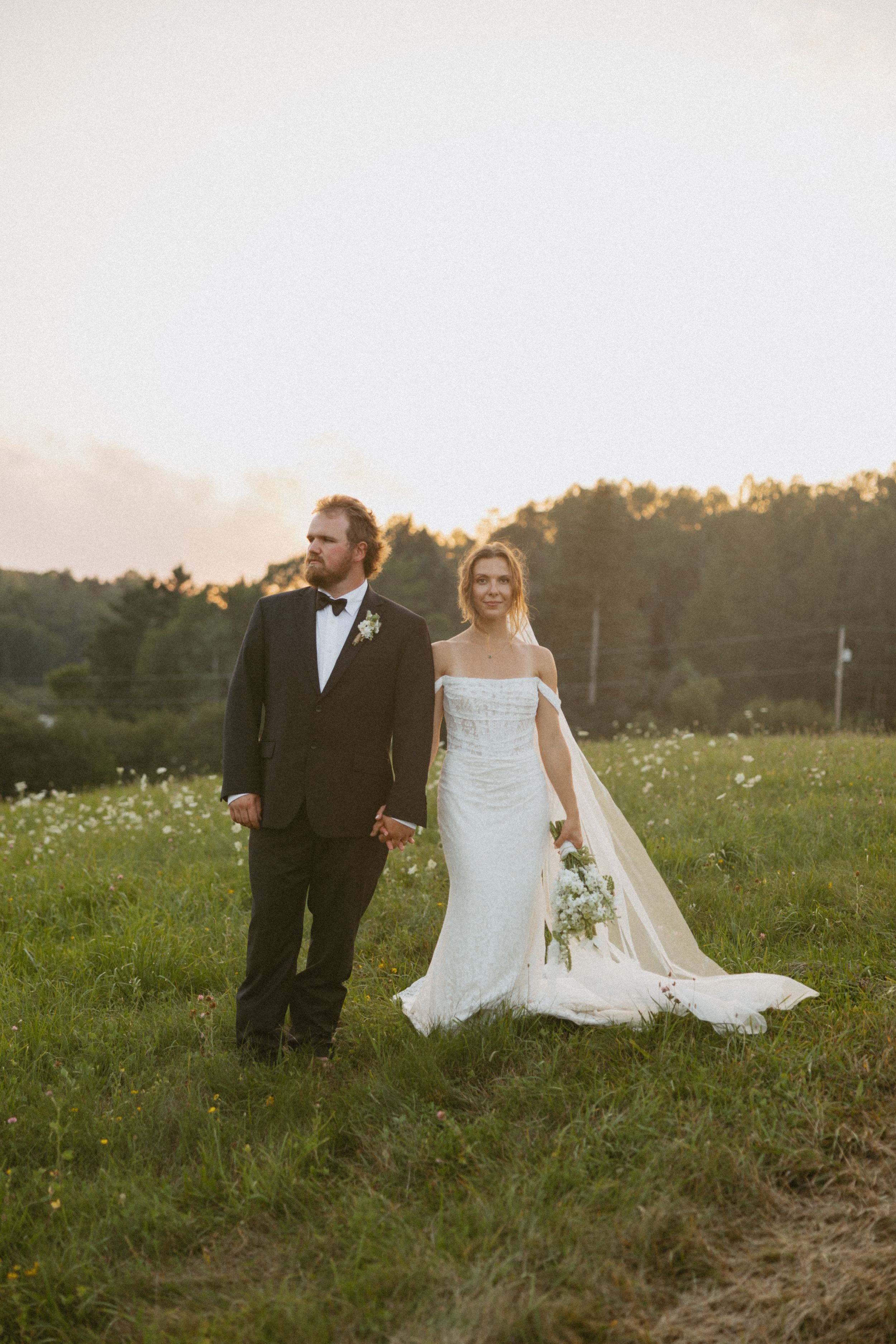 Bride in off-shoulder gown and groom in tuxedo hold hands at golden hour in meadow.