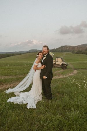 Bride and groom pose together in a lush green field at sunset with rolling hills and a vintage vehicle in the background.