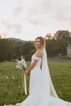 Bride in off-shoulder lace gown with cathedral veil holds white floral bouquet in a sunlit green meadow at golden hour.
