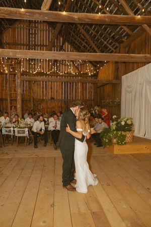 Couple shares their first dance in a rustic barn adorned with string lights while wedding guests watch from decorated tables.