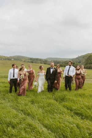 Wedding party walking through lush green meadow with rolling hills, bride in white gown, bridesmaids in dusty rose dresses.
