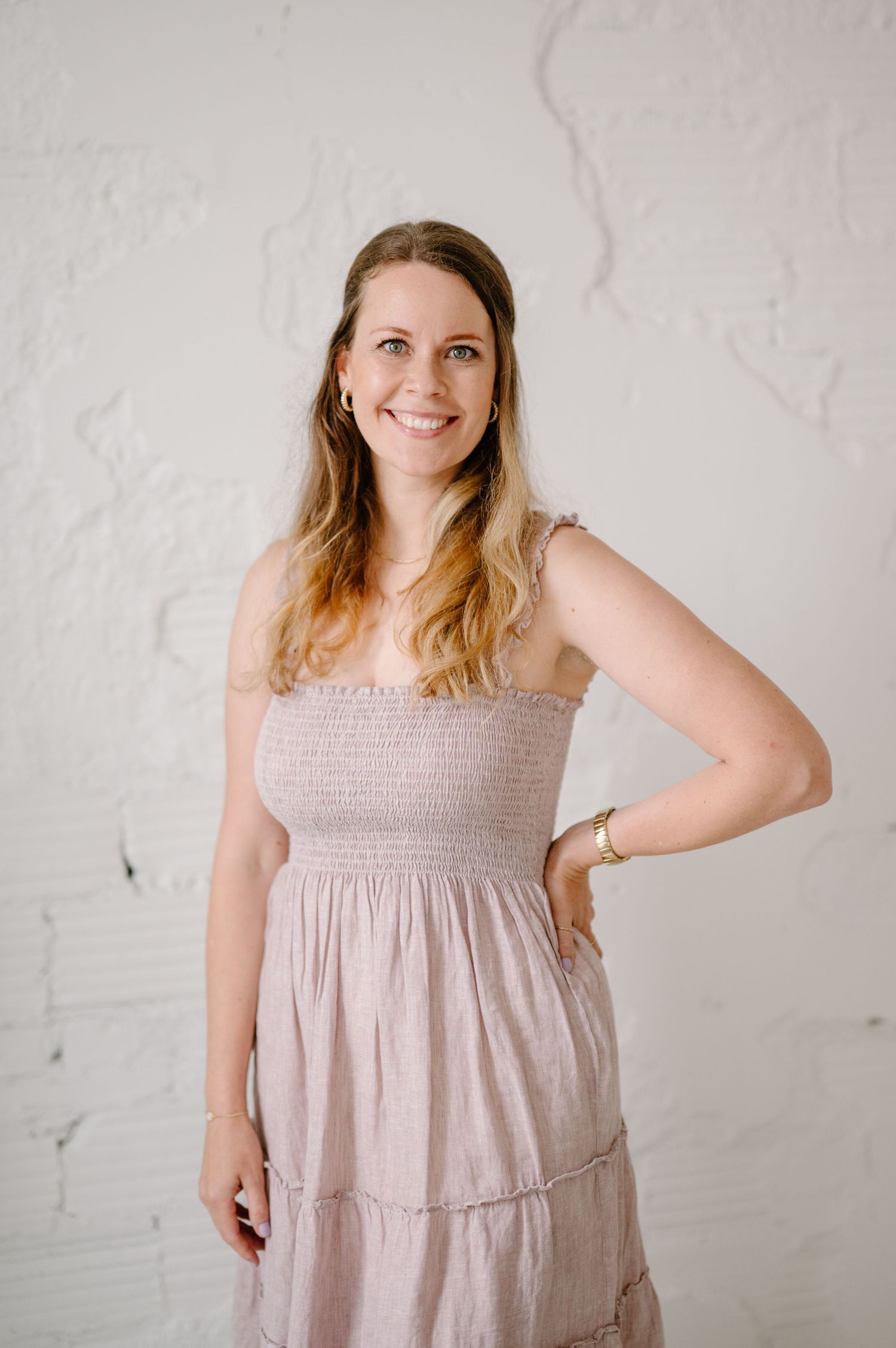 Woman in pink smocked sundress posing confidently against white textured wall, smiling warmly with hand on hip.