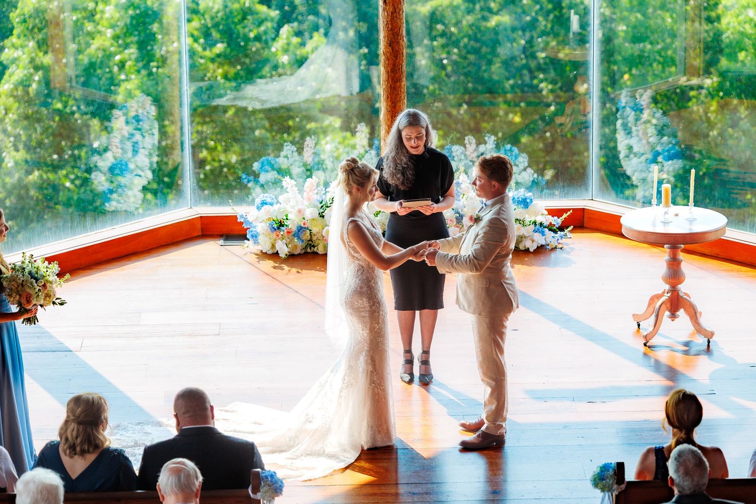 Two brides hold hands during an intimate wedding ceremony in a sunlit glass venue with lush greenery backdrop.