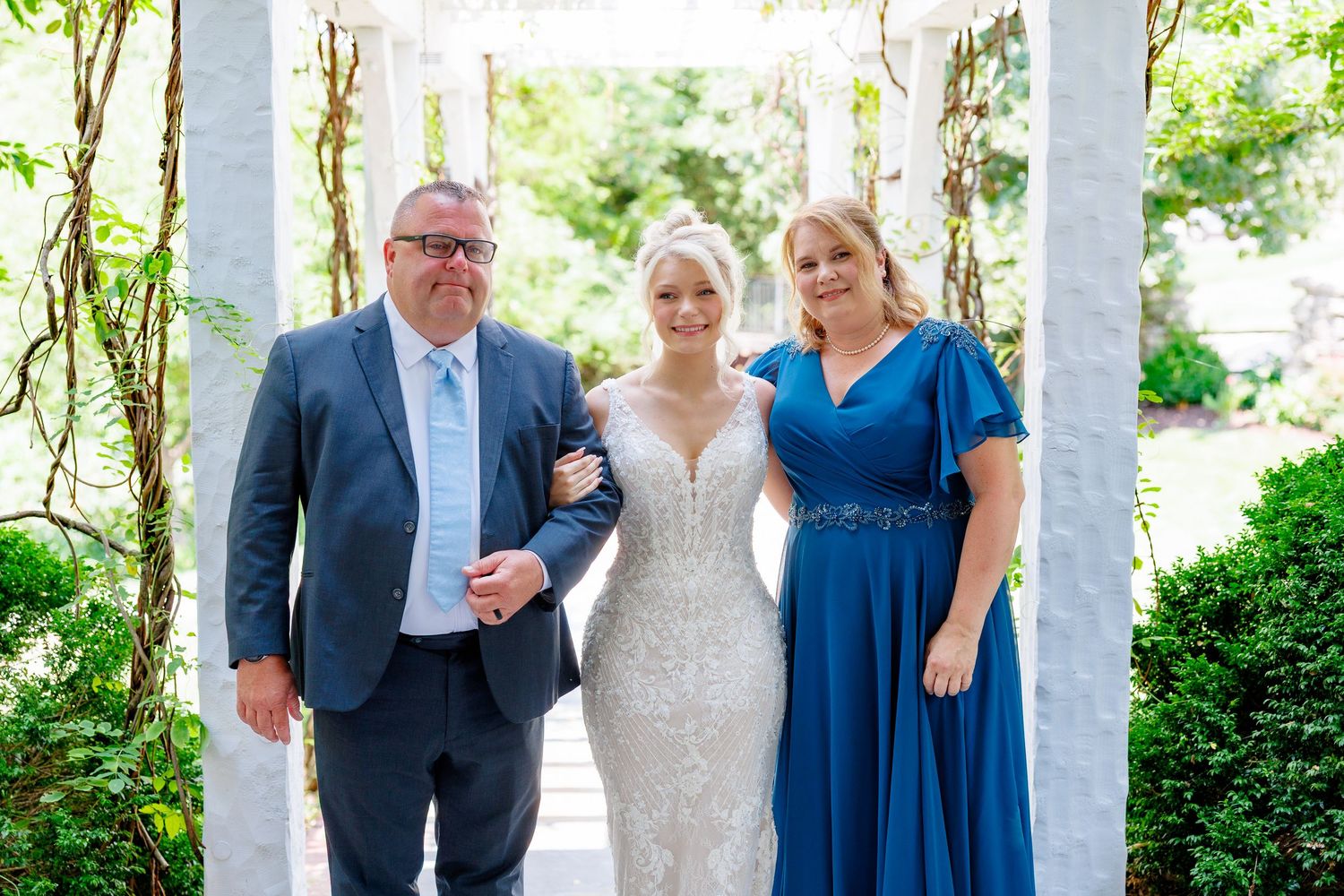 Bride in lace gown poses with man in blue suit and woman in blue dress under white gazebo in garden.