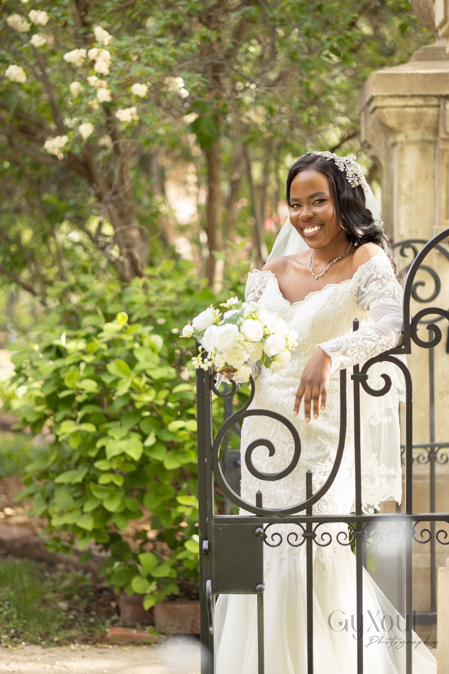 Smiling Black bride in white lace gown holds bouquet by ornate iron gate surrounded by lush greenery.