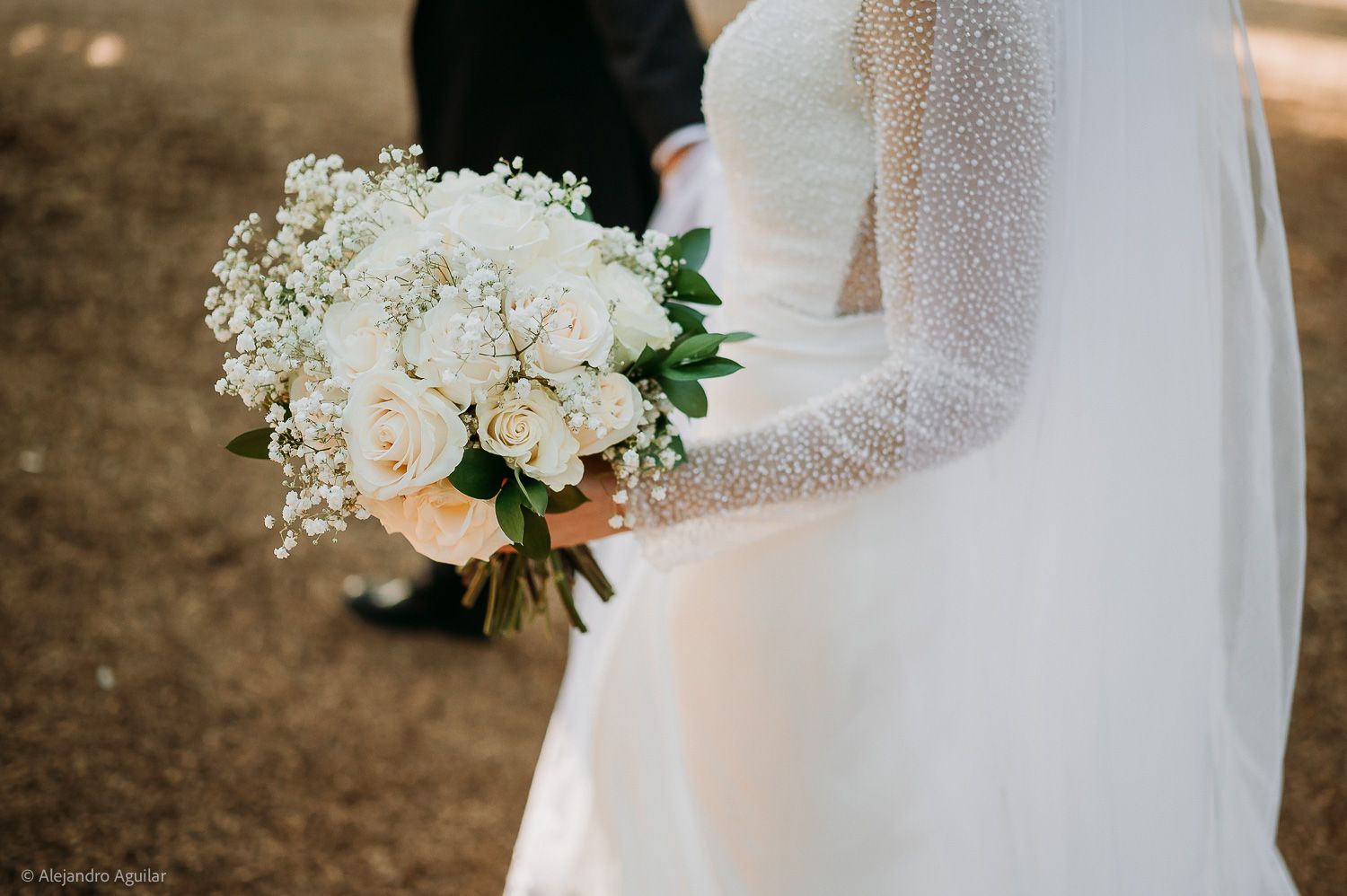 Bride holding a white wedding bouquet of roses and baby's breath, wearing a pearl-embellished long sleeve gown.