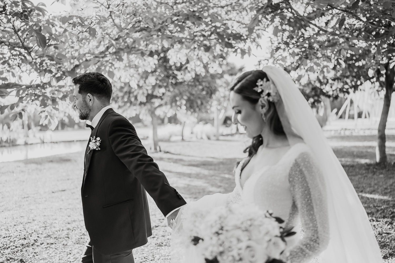 Black and white photo of a bride and groom holding hands walking through a tree-lined garden on their wedding day.