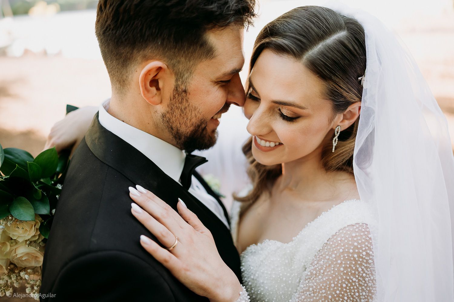 A bride and groom share an intimate moment, touching foreheads, dressed in wedding attire with veil and tuxedo.