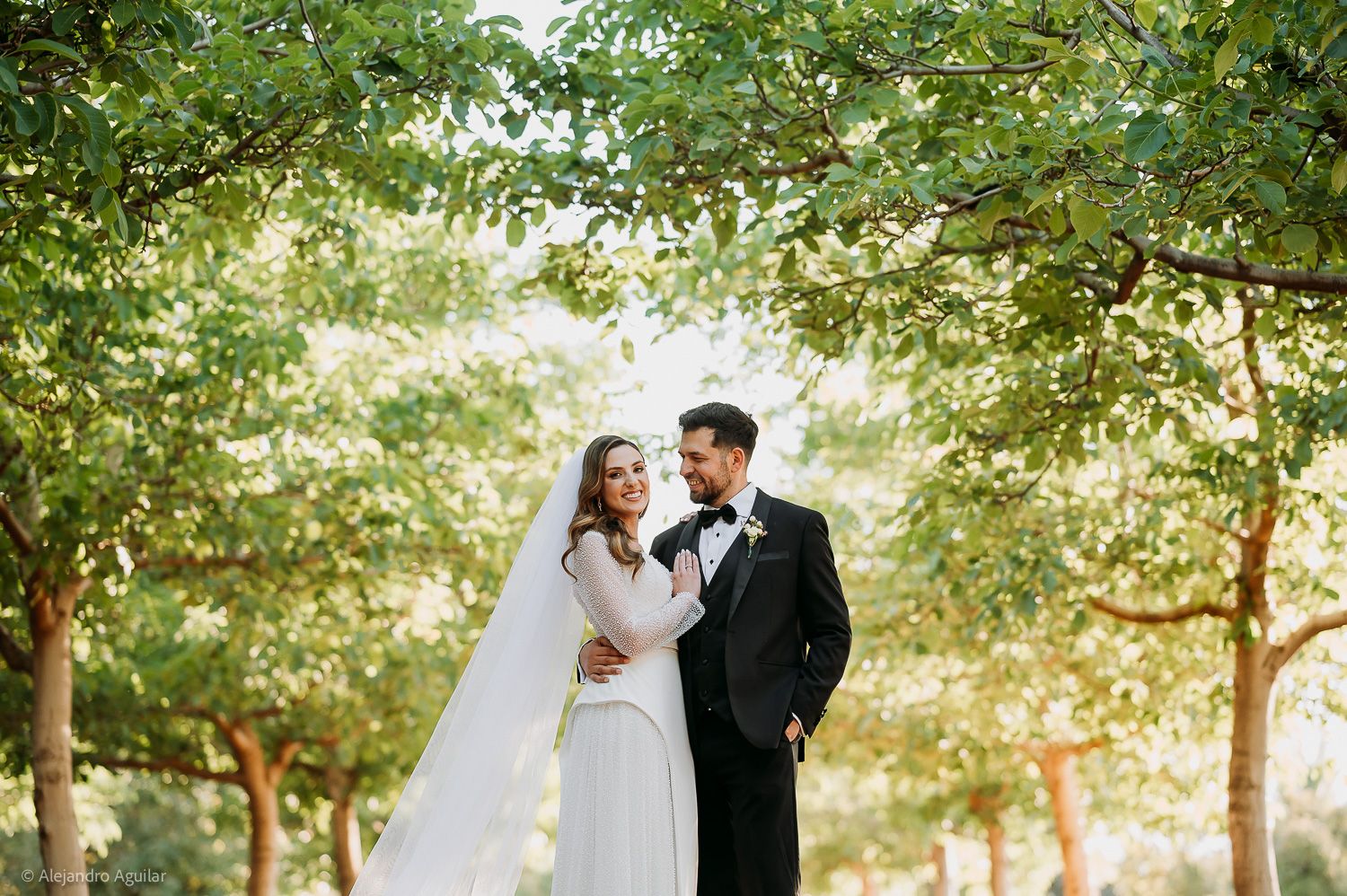 Bride and groom share a romantic moment surrounded by lush green trees in an outdoor wedding portrait.