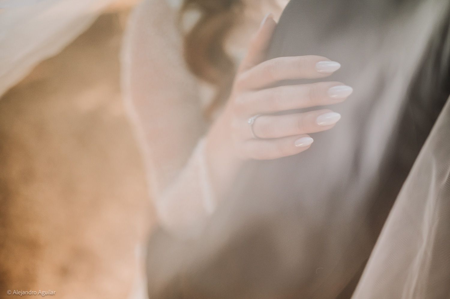 Close-up of a bride's hand with elegant white nails resting on a groom's chest, soft wedding veil in background.