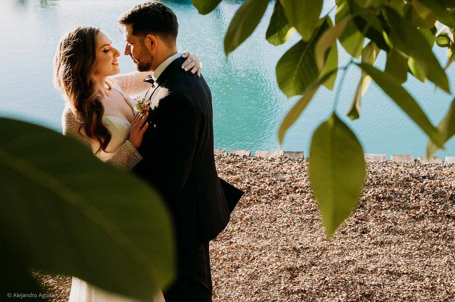A couple in wedding attire embraces near a turquoise lake shore, framed by lush green leaves.