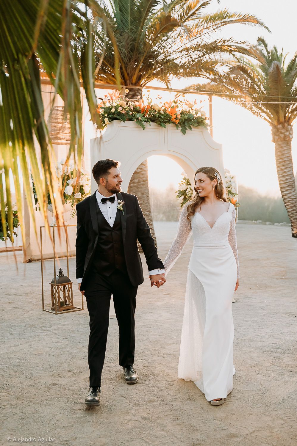 Bride and groom holding hands walking under floral arch with palm trees at golden hour outdoor wedding.