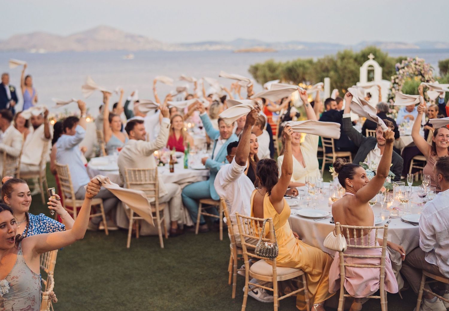Outdoor wedding reception by the sea with guests raising hands in celebration at elegantly set tables under clear skies.