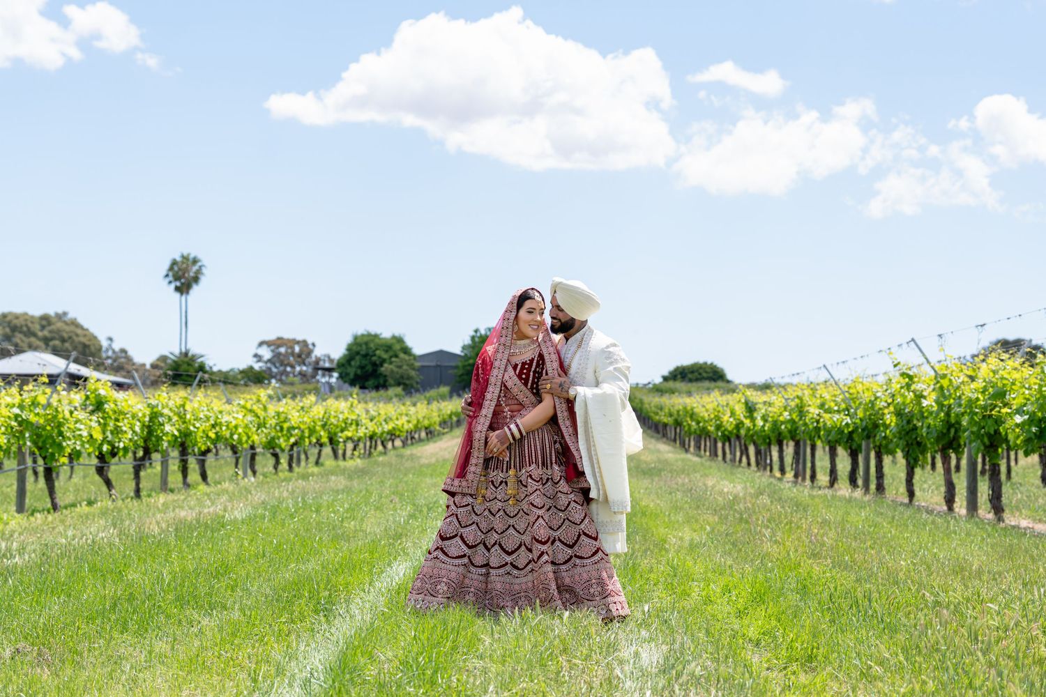 Sikh bride in red lehenga and groom in white sherwani holding hands outdoors on a sunny day.