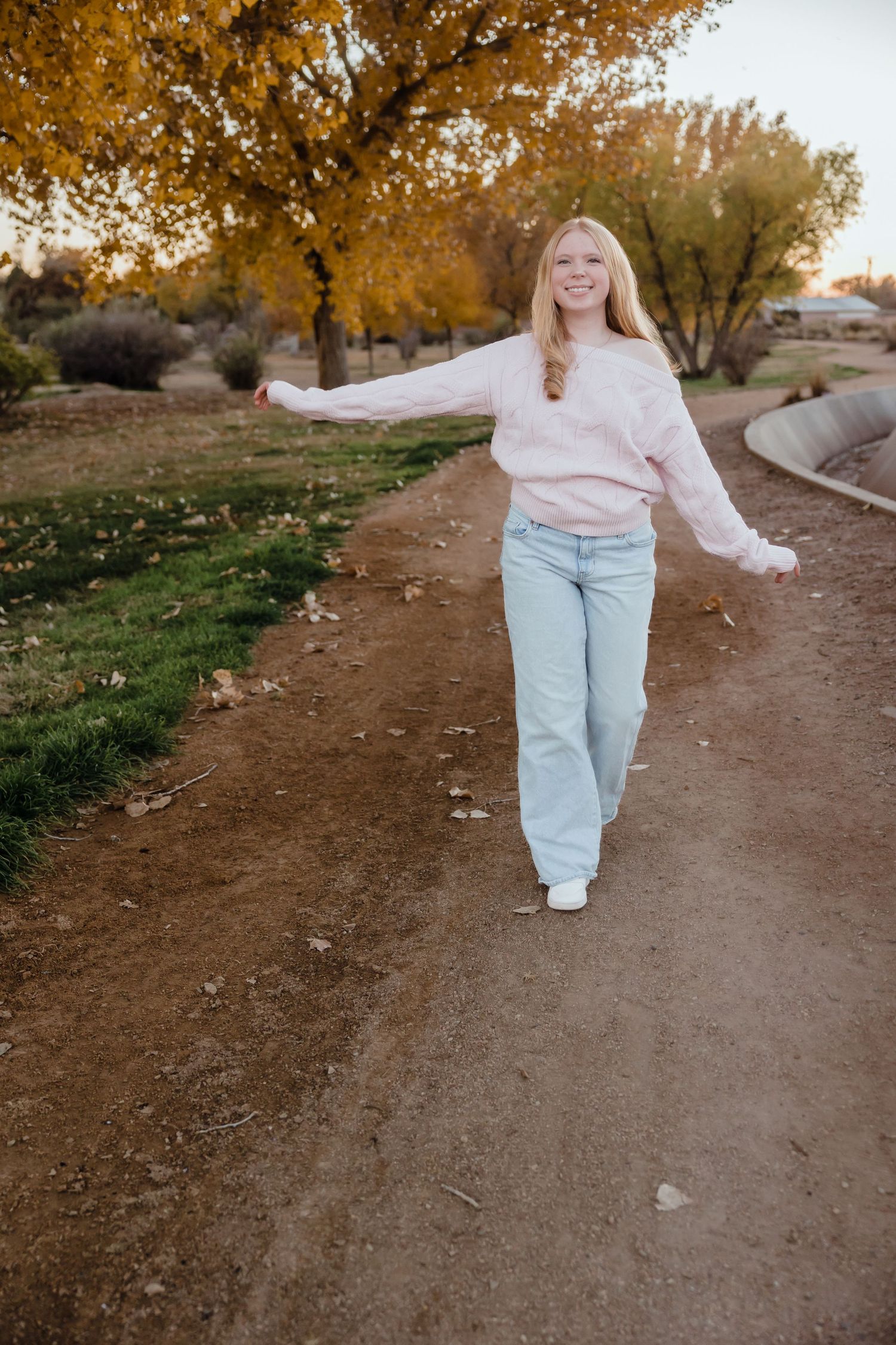 Young woman in pink sweater and jeans walks along a dirt path surrounded by autumn trees.
