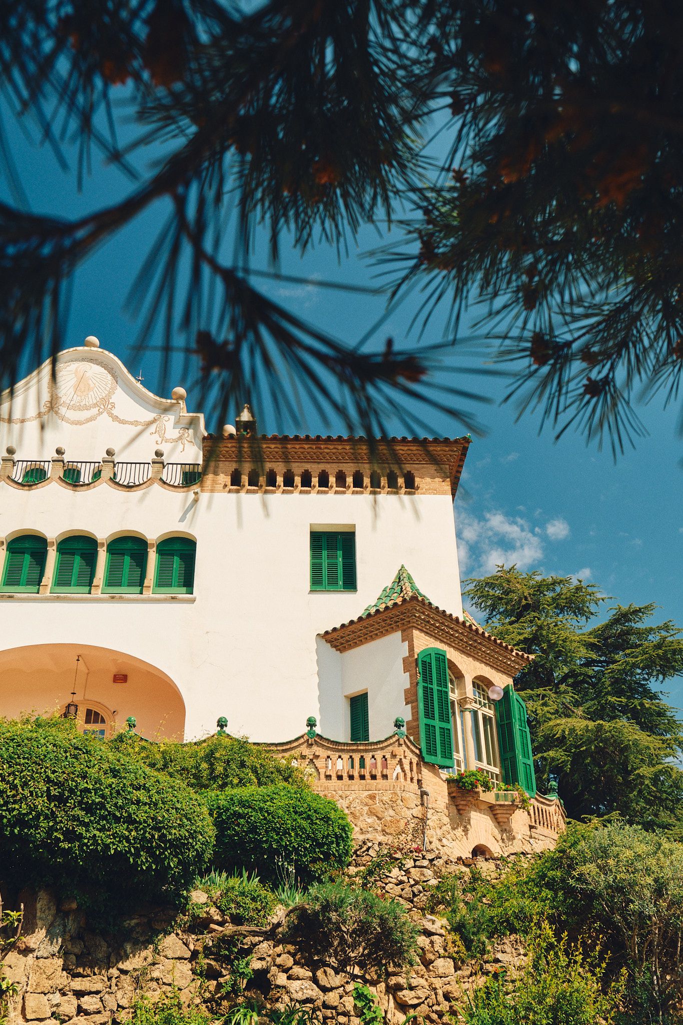 White Mediterranean villa with green shutters and ornate architecture surrounded by lush gardens under a blue sky.