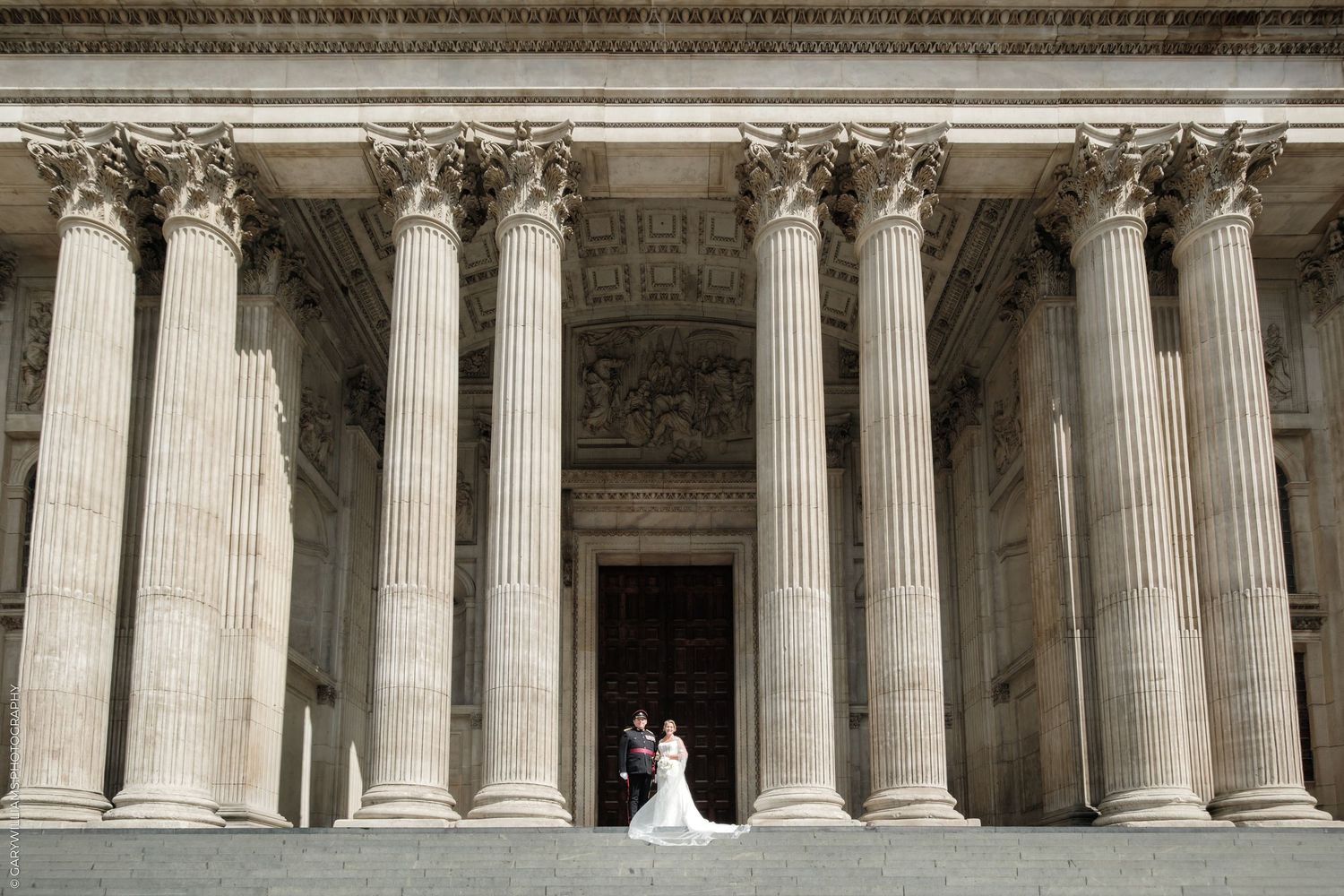 A bride and groom pose before grand Corinthian columns of a neoclassical building on their wedding day.