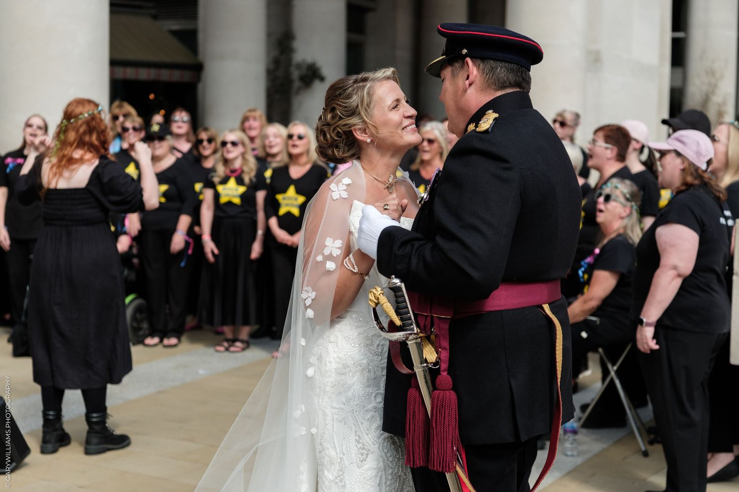 Military groom in tartan kilt embraces bride in white lace wedding gown outdoors with choir in black shirts behind them.