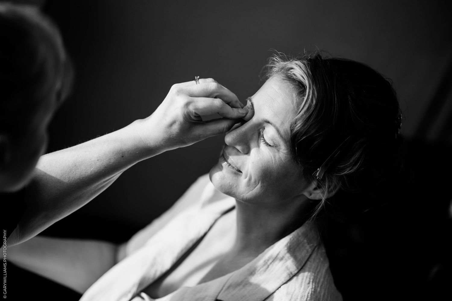 Black and white photo of a woman smiling as a makeup artist applies eyeshadow during bridal preparation.