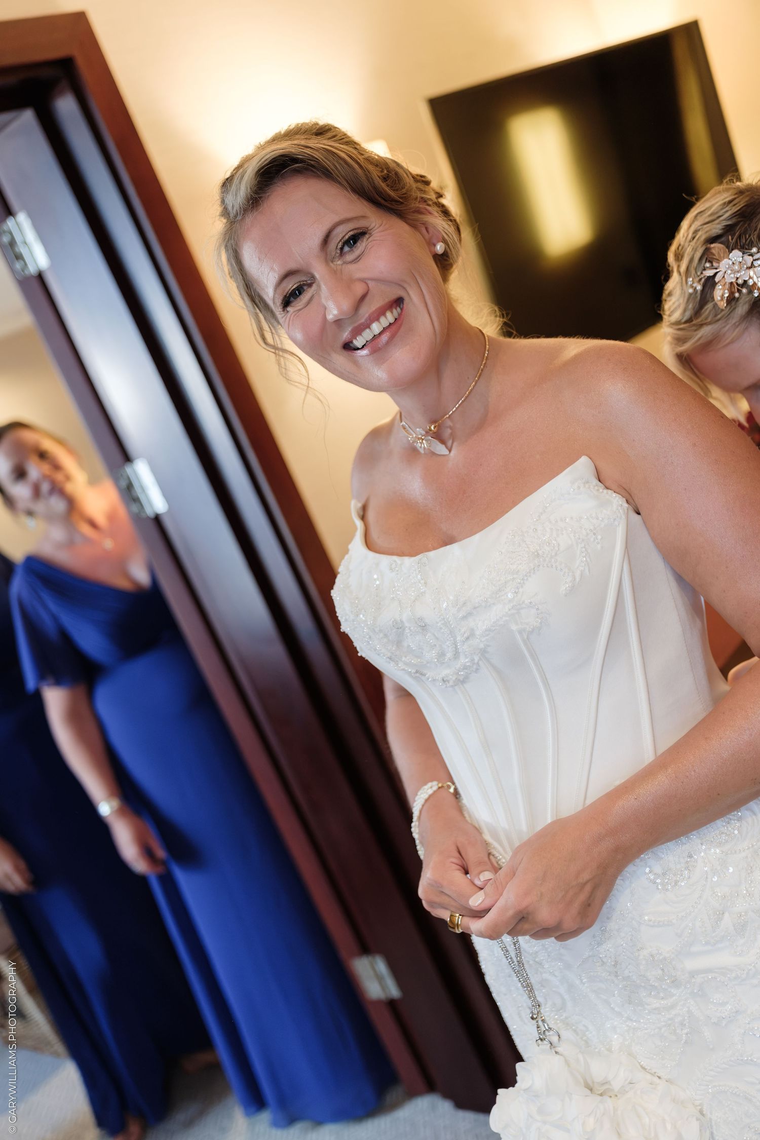 Smiling bride in white strapless lace gown with bridesmaids in blue dresses reflected in mirror during wedding preparations.
