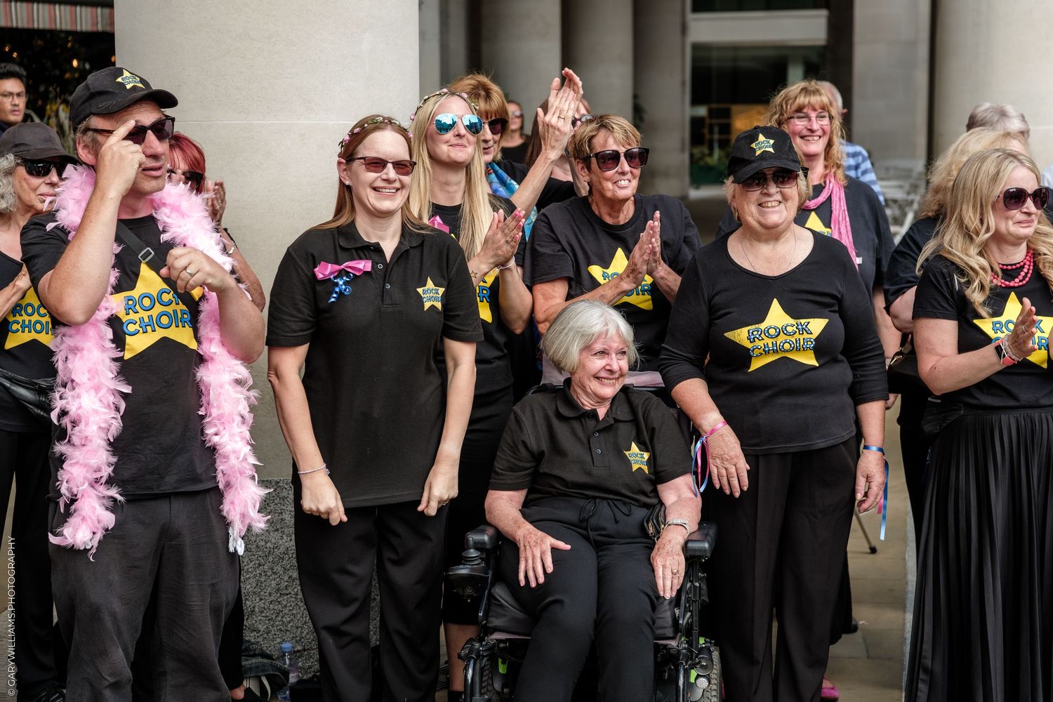 Group of women in matching black Rock Star shirts posing together outdoors, one seated in wheelchair, wearing pink feather boa.