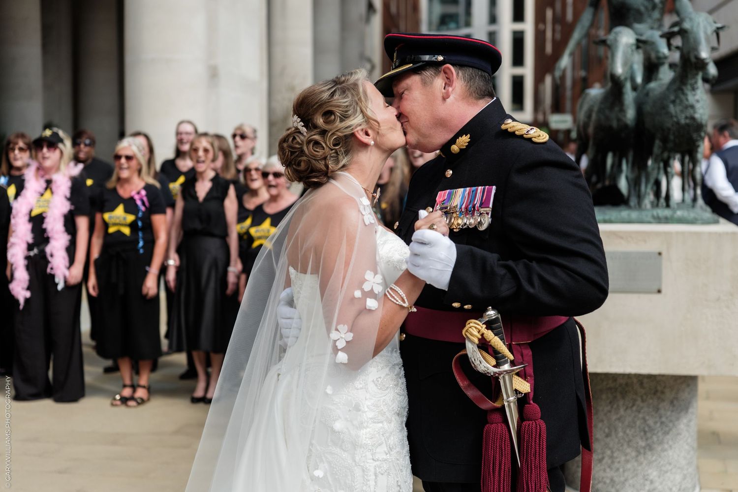 Military groom in uniform kisses bride in white wedding dress and veil outdoors at a formal wedding ceremony.