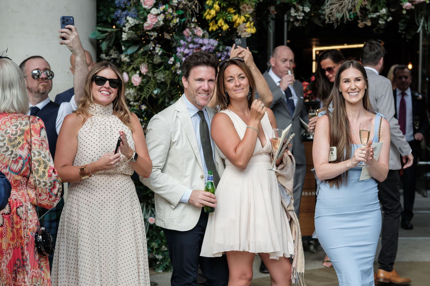 Elegantly dressed guests socializing outdoors at a garden party, holding champagne glasses beneath floral decorations.