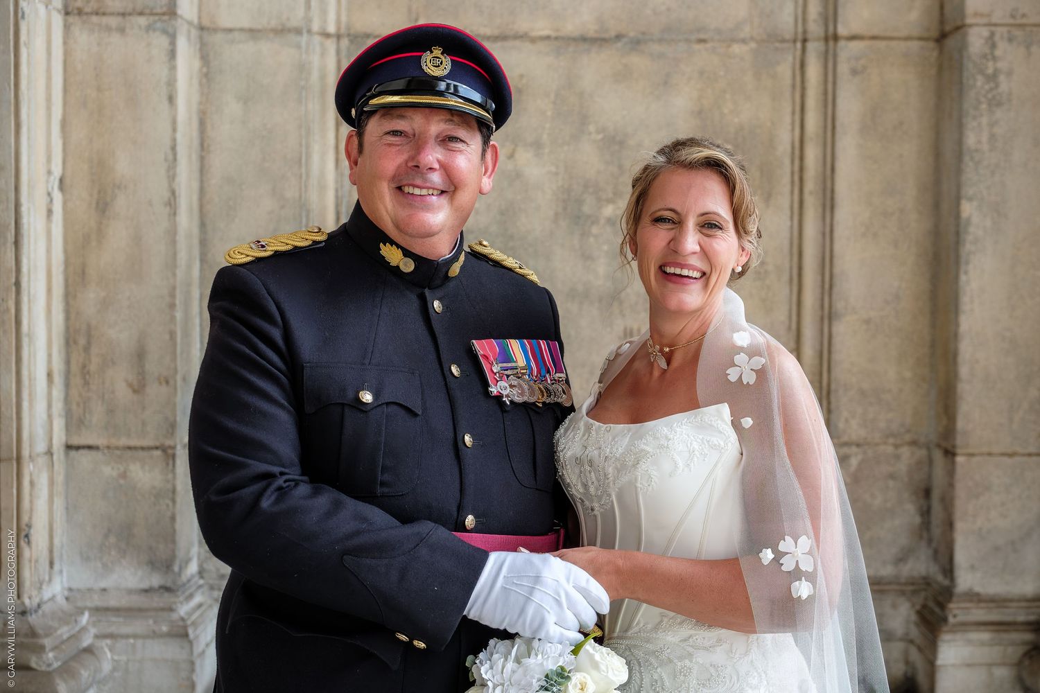 Military groom in dress uniform with medals poses with smiling bride in white gown and veil on wedding day.