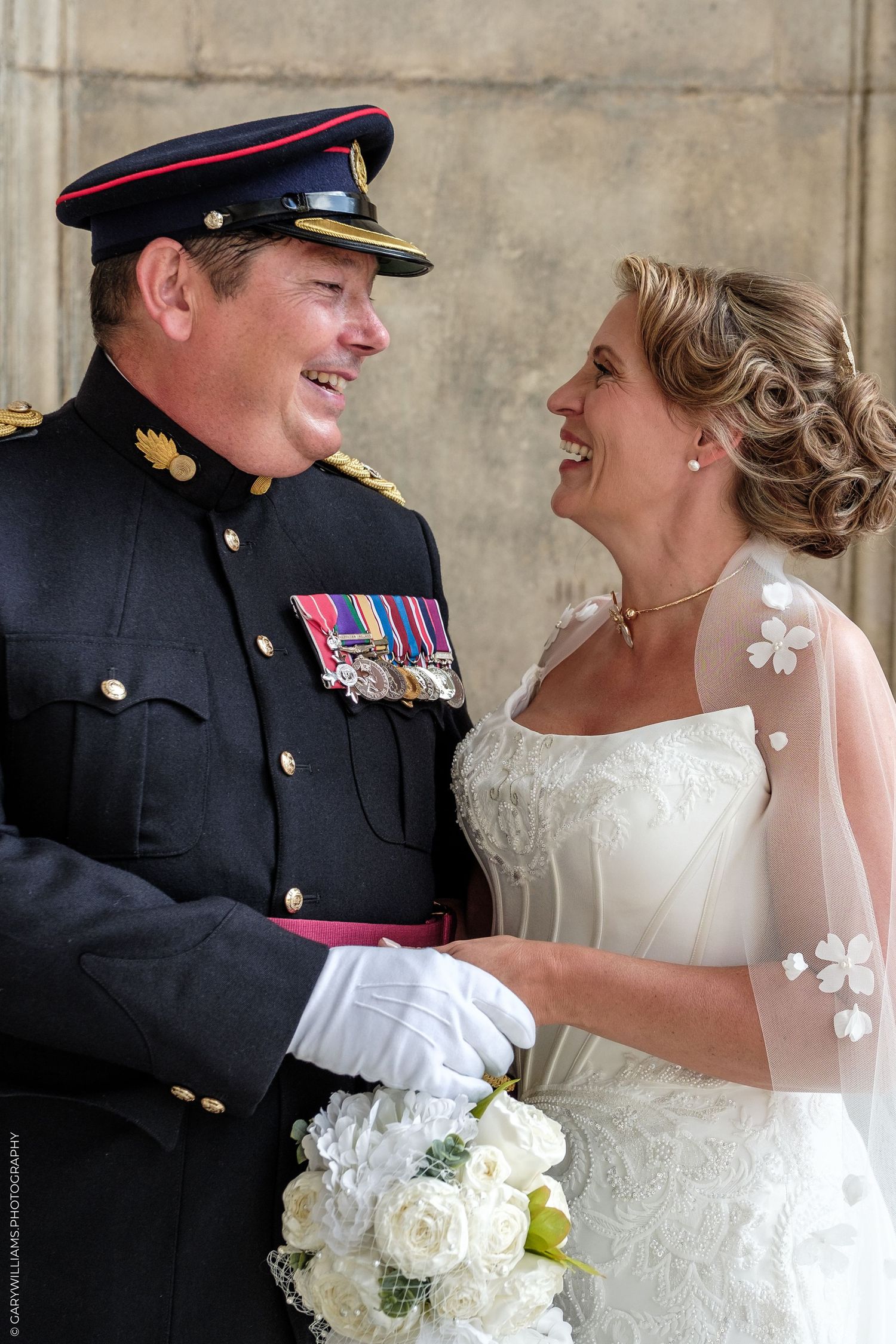 Military groom in uniform with medals laughs with bride in white lace wedding dress holding bouquet of white roses.