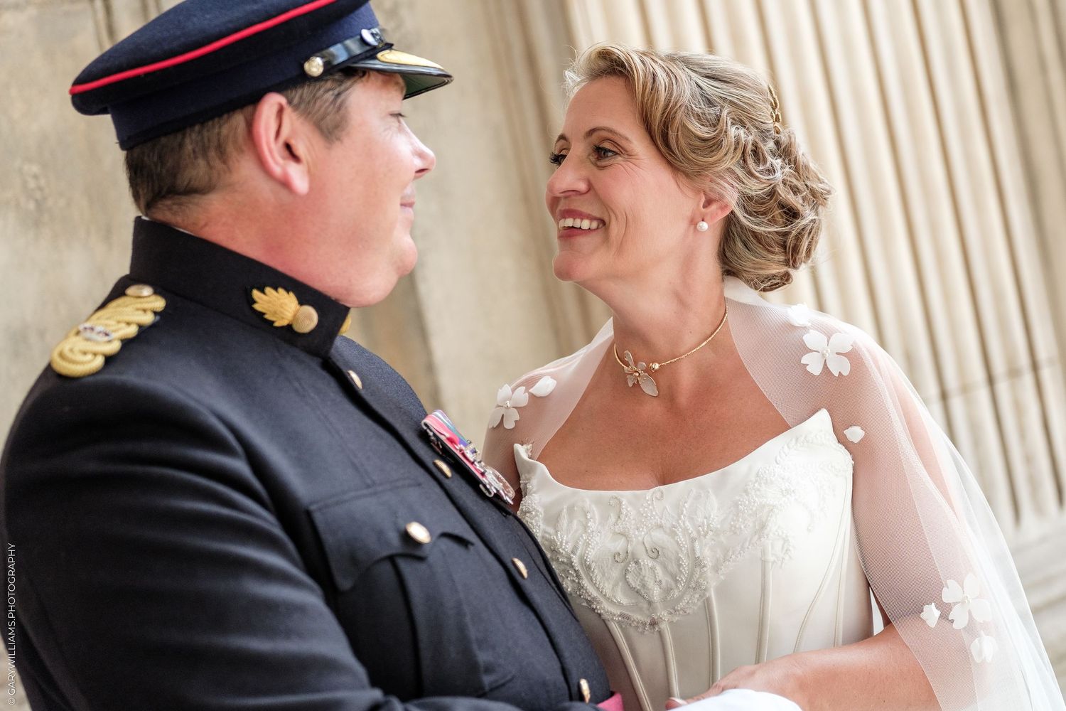 Military groom in uniform smiles at bride in white wedding dress with floral veil during outdoor ceremony.