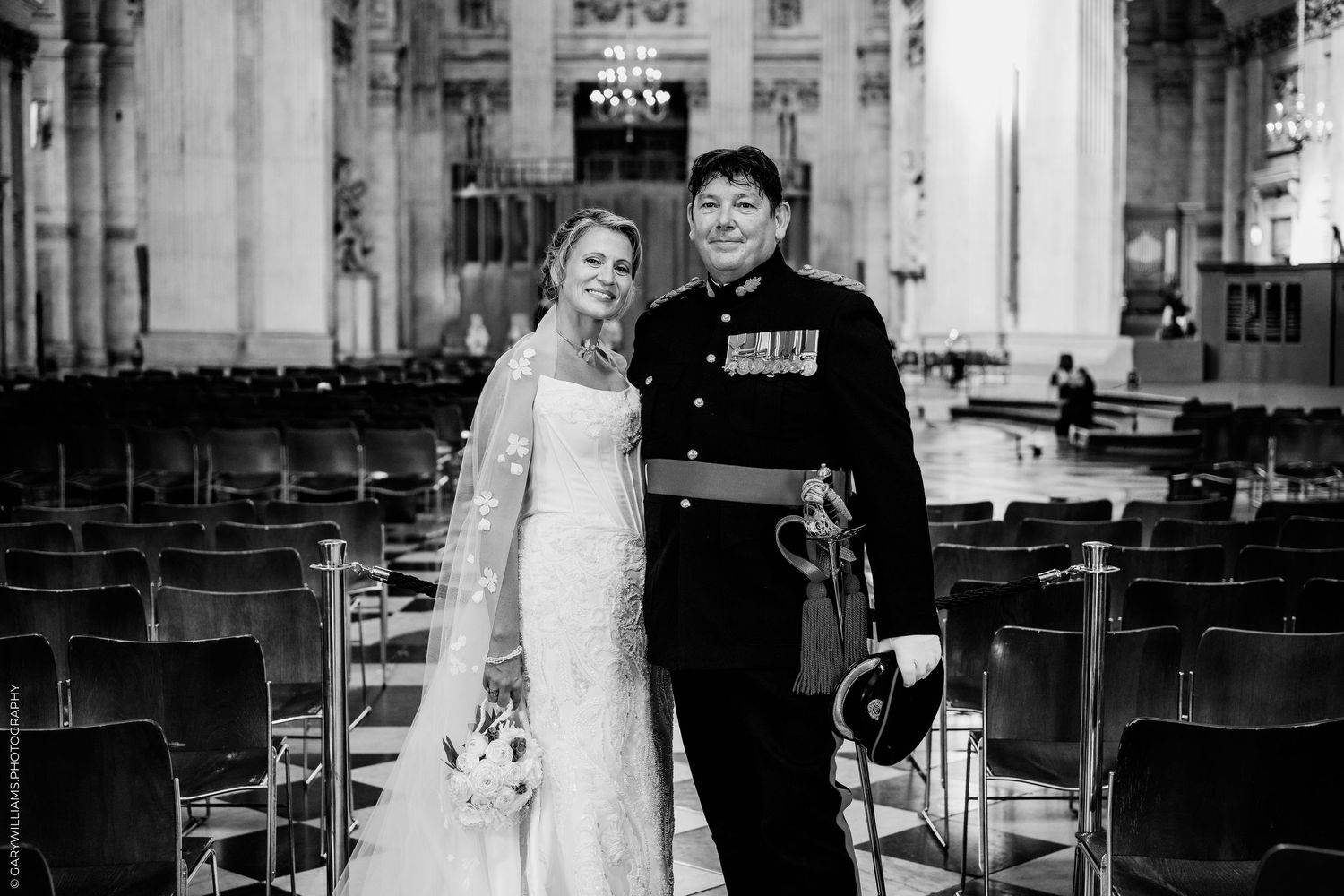 Black and white wedding photo of bride in lace gown and groom in military uniform with sword inside grand cathedral.