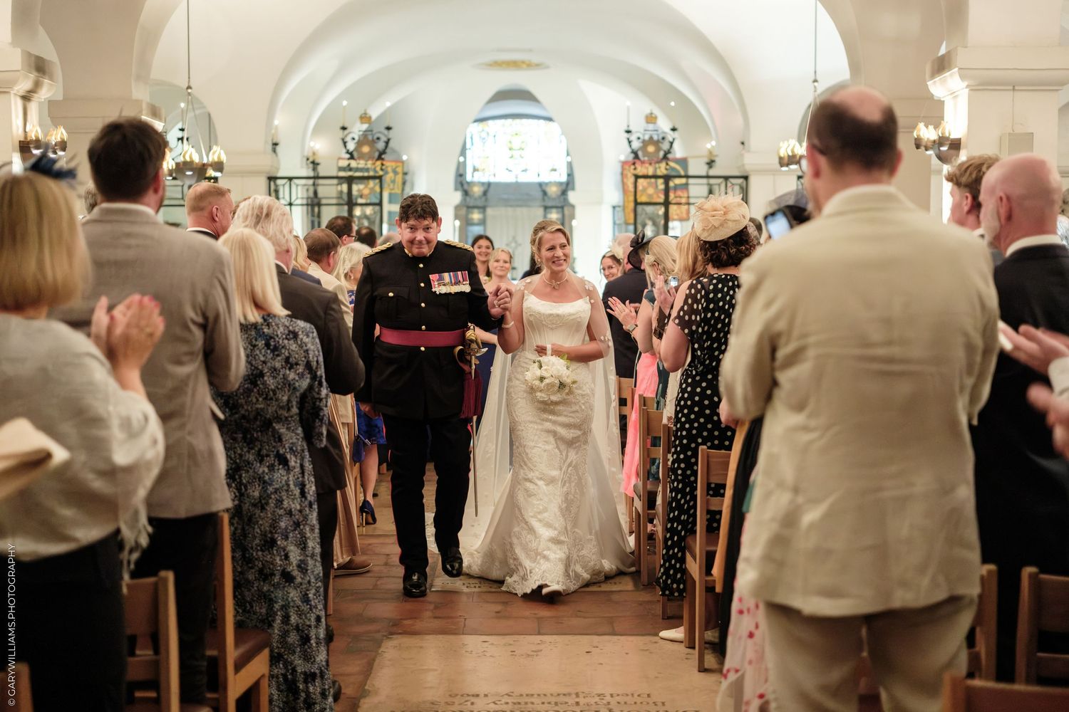Newlywed couple walks down church aisle in wedding ceremony, bride in white gown, groom in military uniform, guests applauding.