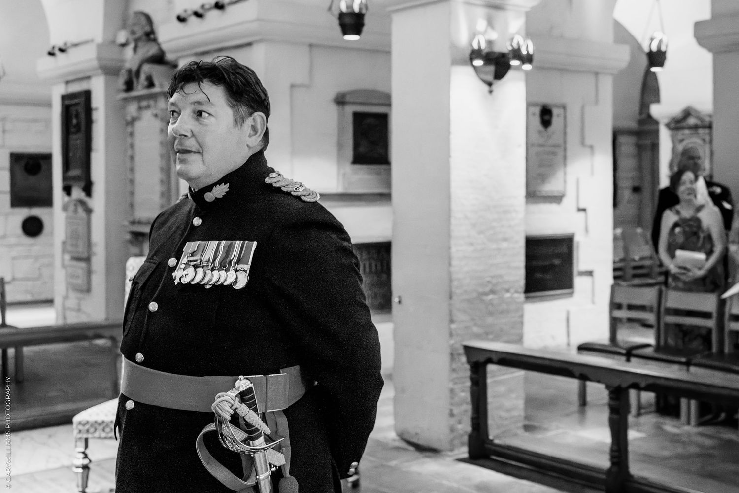 Military officer in dress uniform with medals stands in historic church interior, black and white photography.