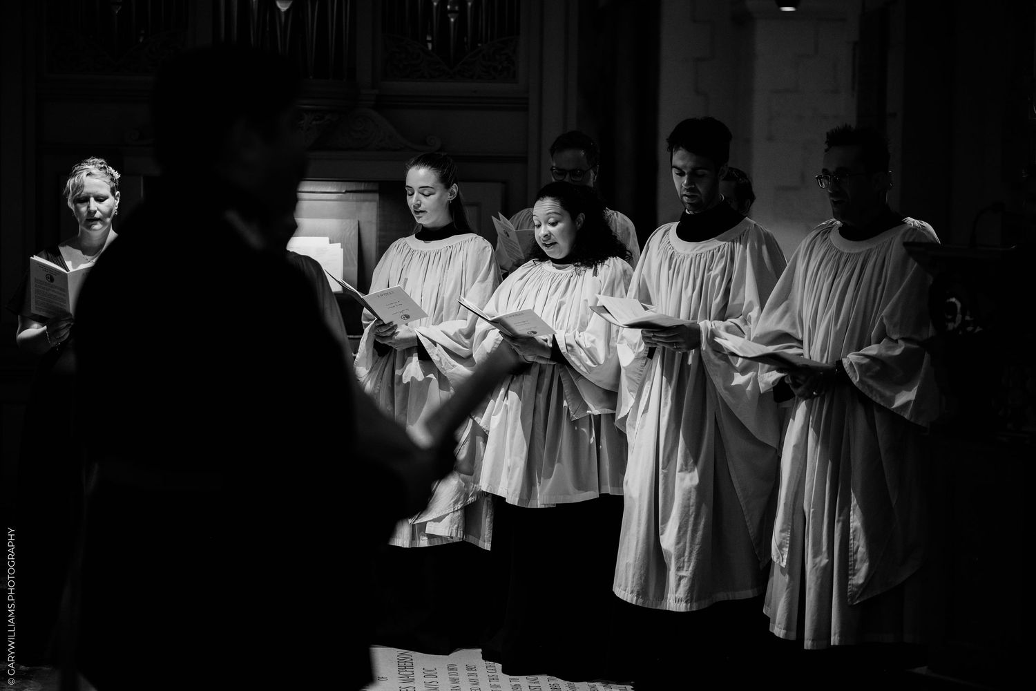 Black and white photo of choir members in white robes singing from sheet music in a church.