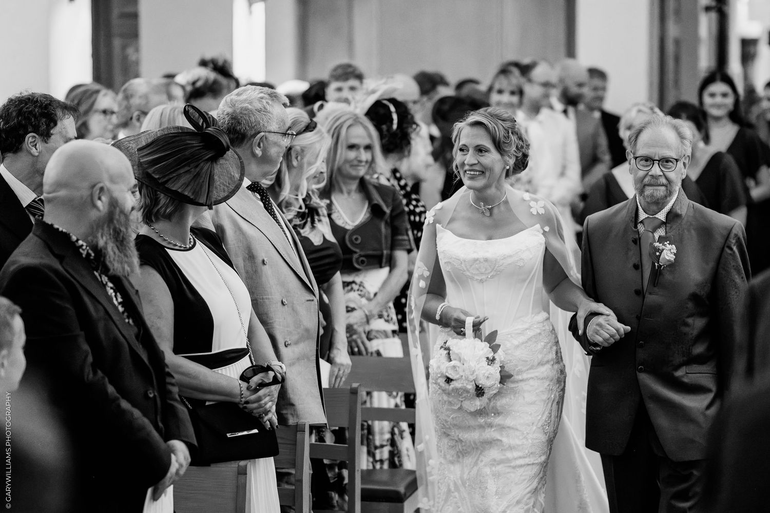 Bride walks down the aisle smiling with bouquet during wedding ceremony in black and white photo.