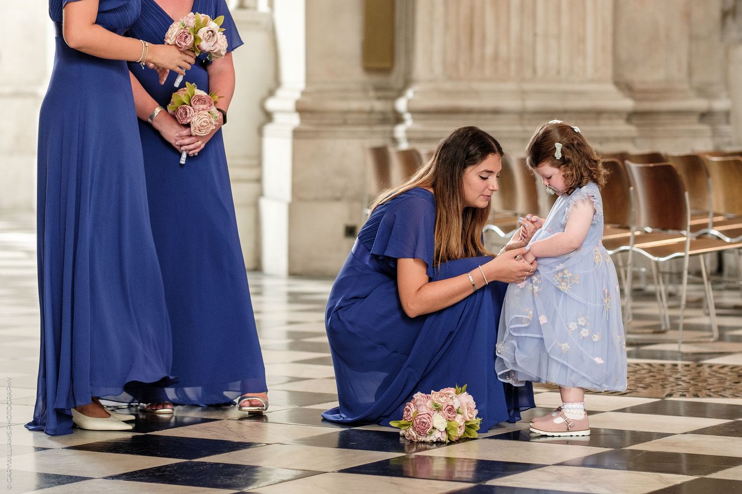 Bridesmaid in blue dress kneeling to tend to flower girl inside a grand cathedral wedding venue.