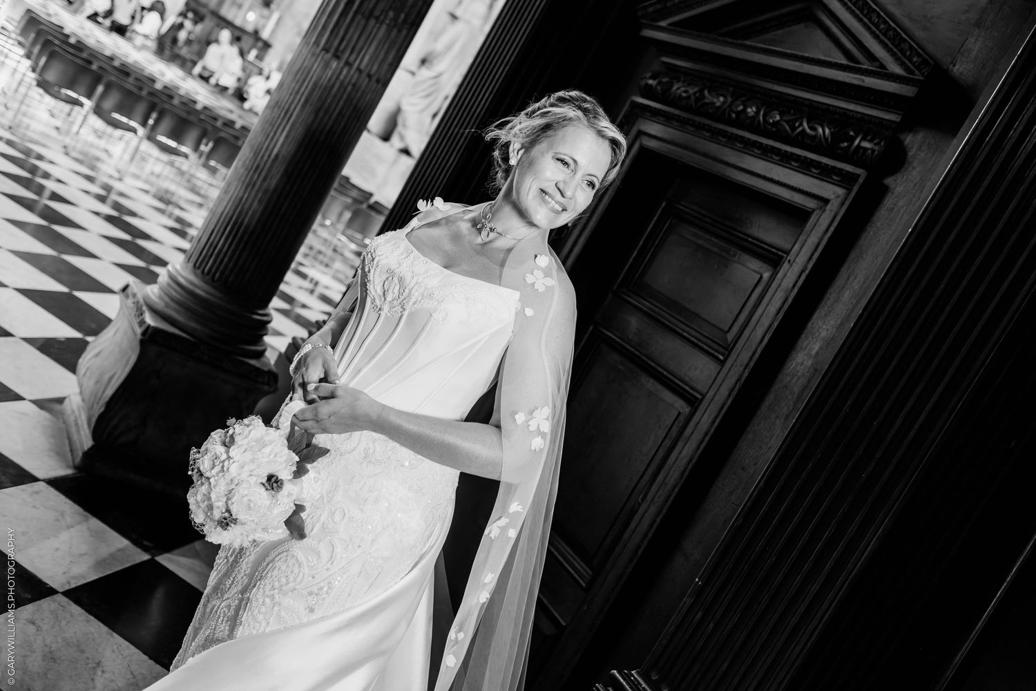 Elegant bride in lace wedding dress holding bouquet, posing by ornate door in black and white photo.