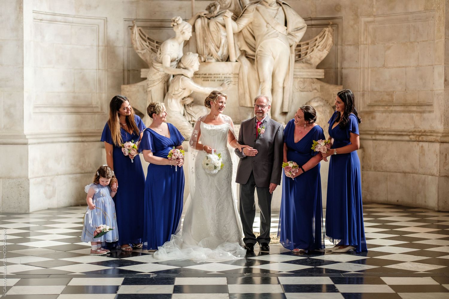 Wedding party poses before marble sculpture on checkered floor, bride in white gown with bridesmaids in royal blue dresses.