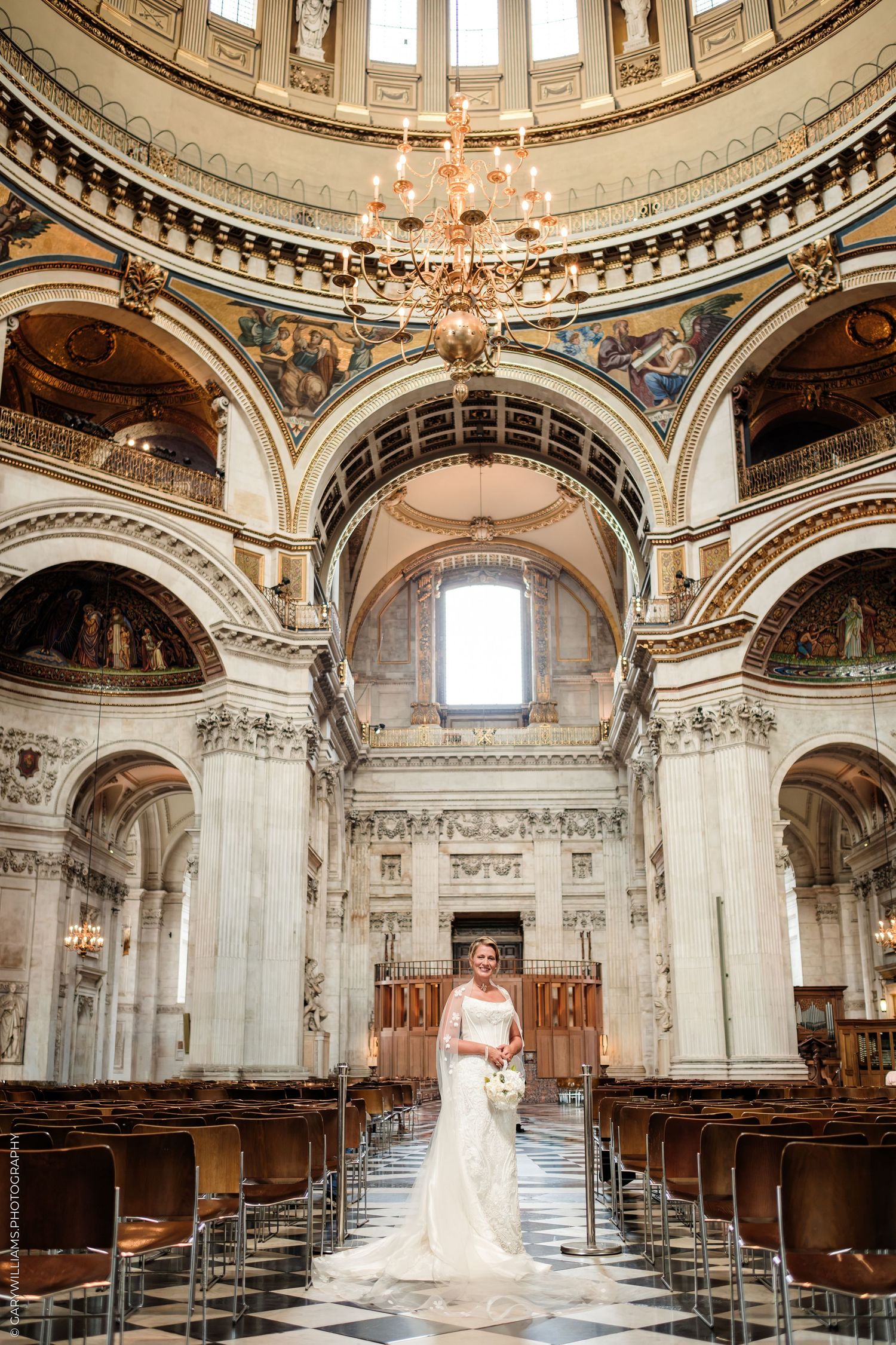 Bride in white gown standing in the grand aisle of St. Paul's Cathedral, London, with ornate architecture and chandelier.