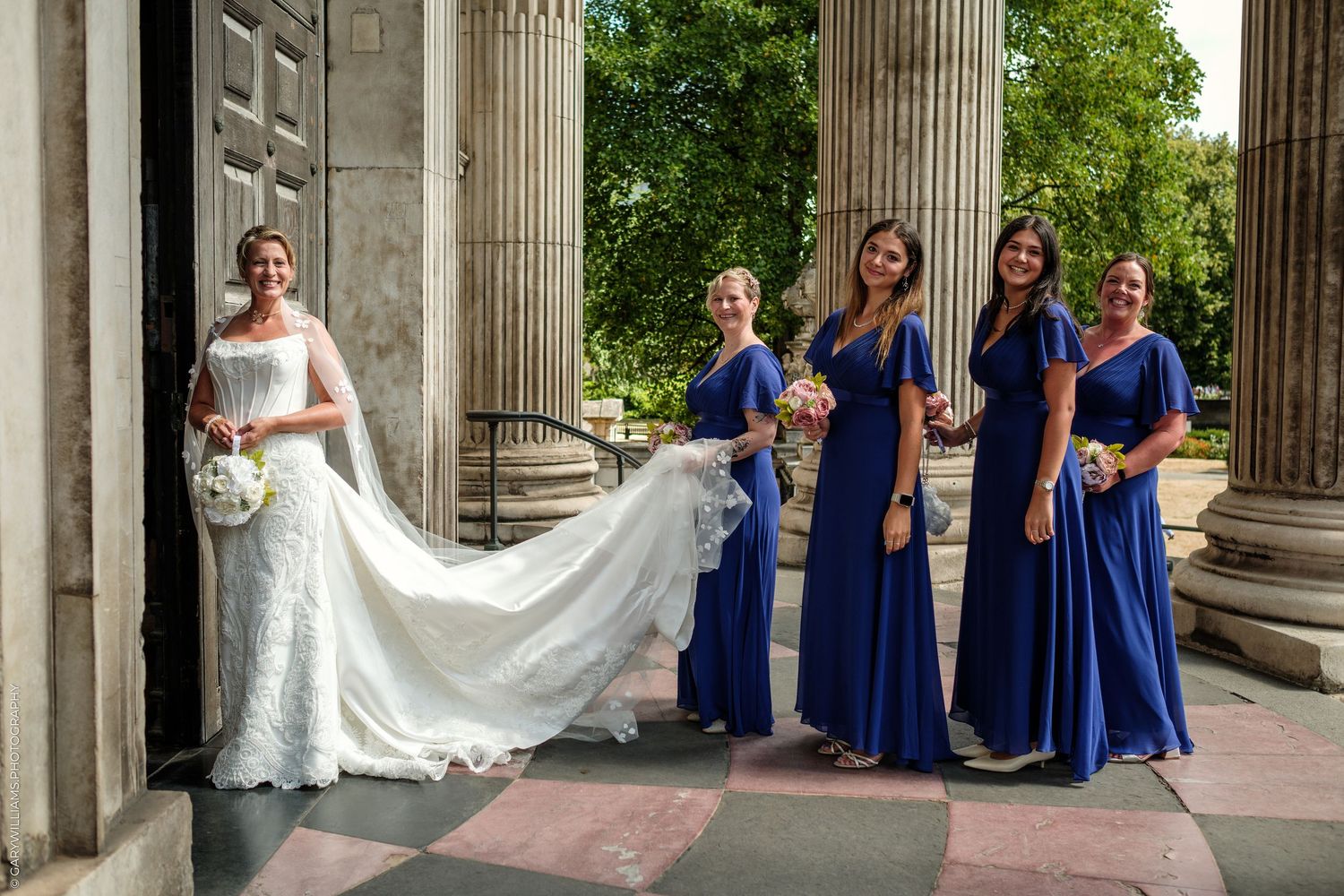 Bride in white gown with bridesmaids in royal blue dresses posing at elegant columned building entrance.